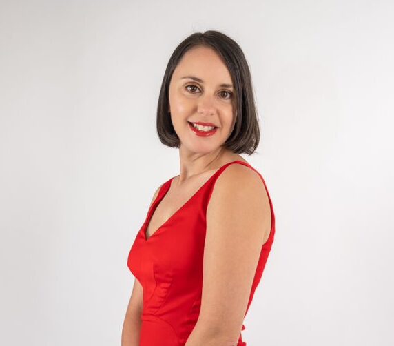 Image of Jessica Zammit. Jessica is standing side on, with short brown hair. She is smiling into the camera and wears a red dress. 